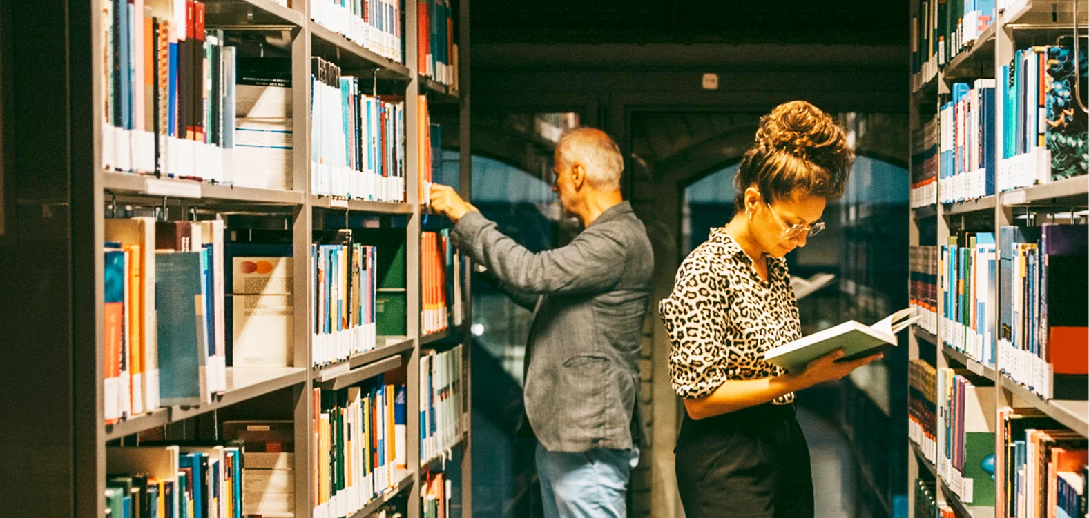 A man and a woman stand in a library looking at books on the shelves.