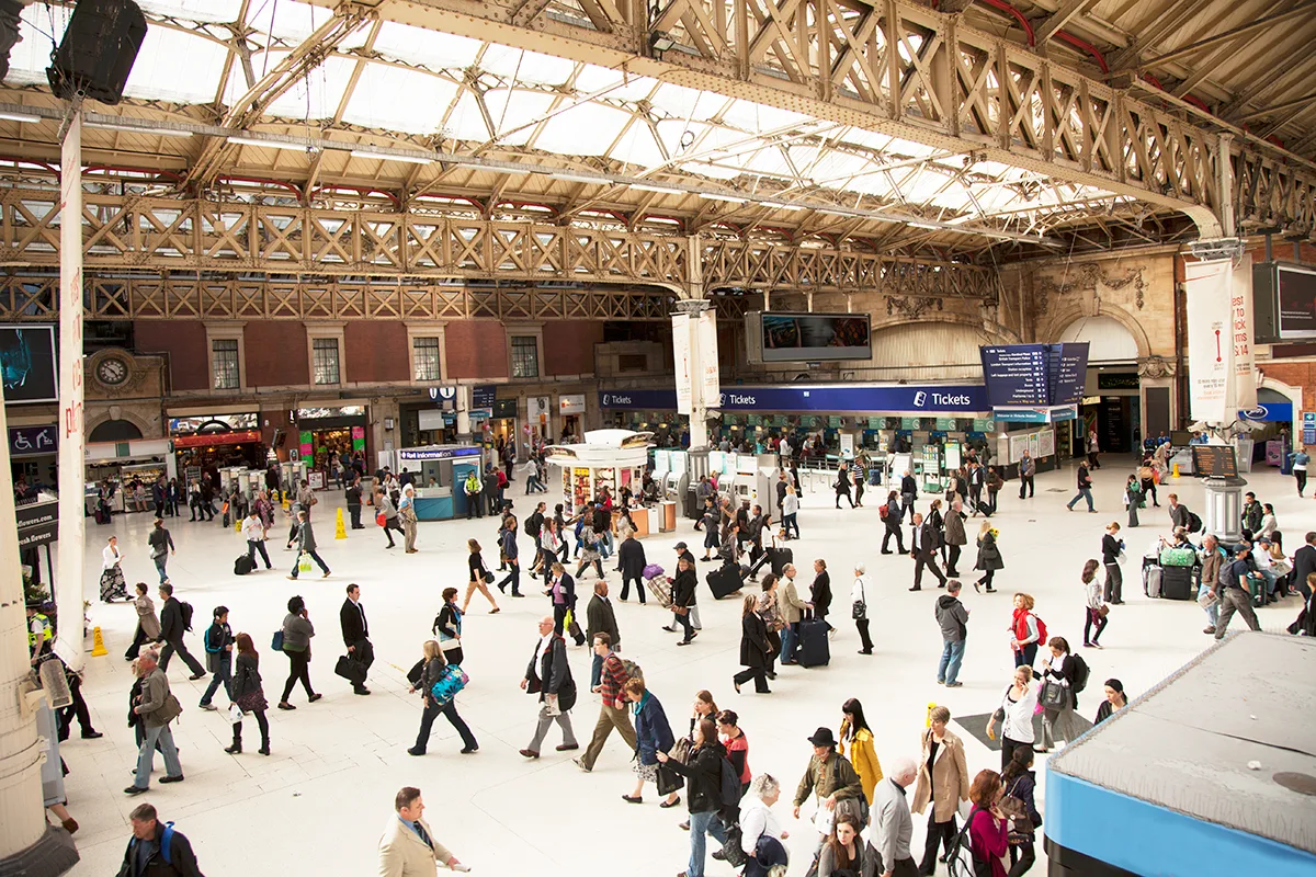Concourse at London Victoria station with commuters walking across the white floor beneath ornate metal roof structure, and shops and ticket counters to the sides.