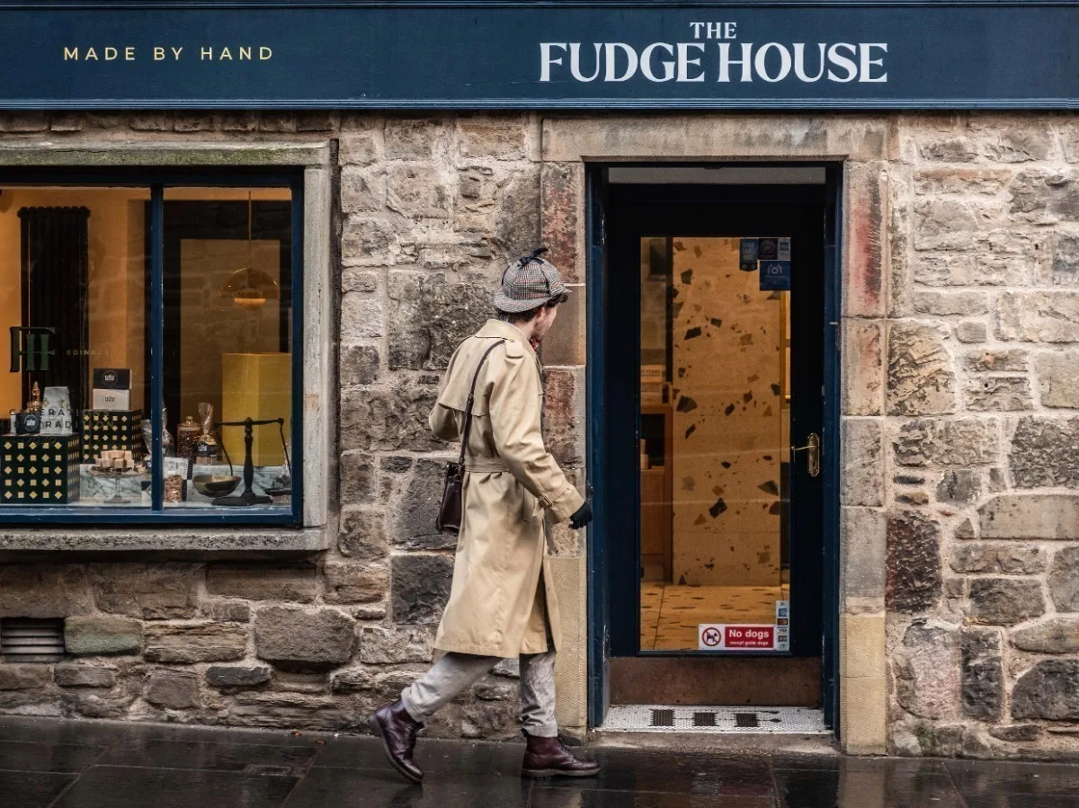 A white man dressed as Sherlock Holmes with a long tan raincoat and deerstalker hat, approaching the door of a shop.