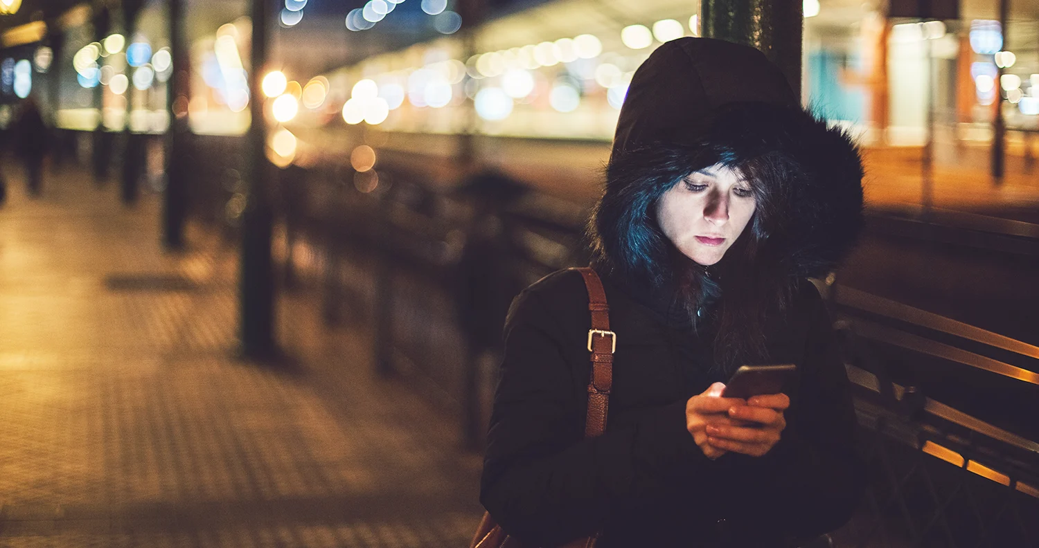 A woman standing alone at night, looking down at her mobile phone.