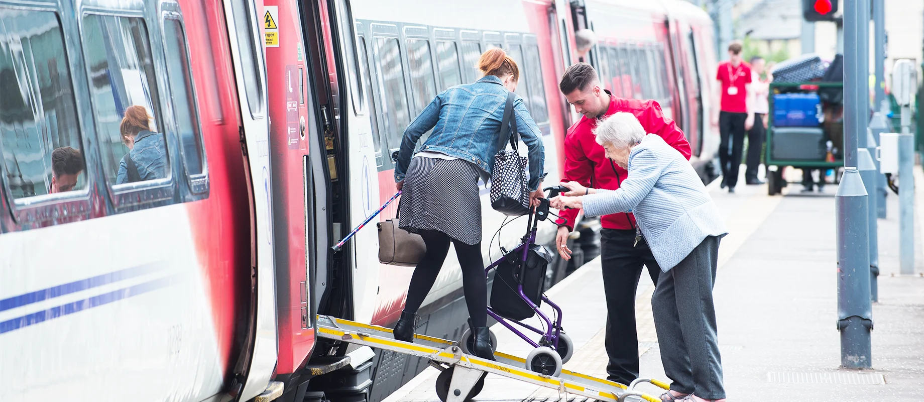 A railway employee helps an elderly woman using a wheeled walker up a ramp onto a train at a station platform, while a passenger guides the walker.