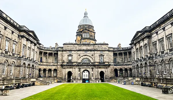An old stone university building with a dome on one, side, with a lawn in the centre of a courtyard. 