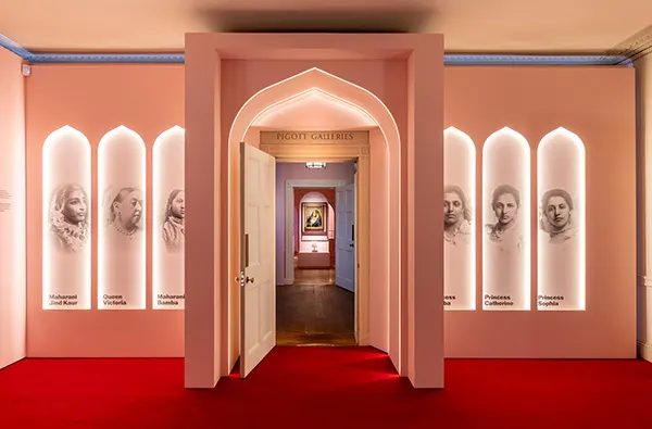 Pink gallery entrance with arched doorway, red carpet, and illuminated portrait panels of historical figures including Queen Victoria.