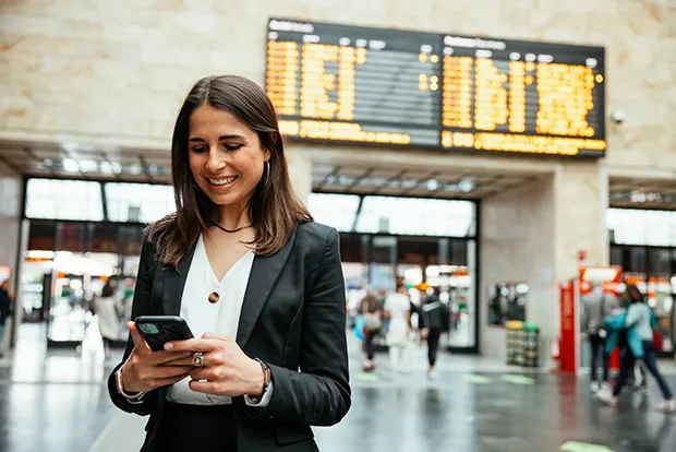 A woman with long brown hair and smiling, checks her phone in a railway station concourse.