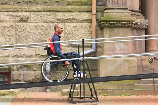 A smiling young Black man with cropped hair, wheels his wheelchair up a ramp outside an old, stone building. 