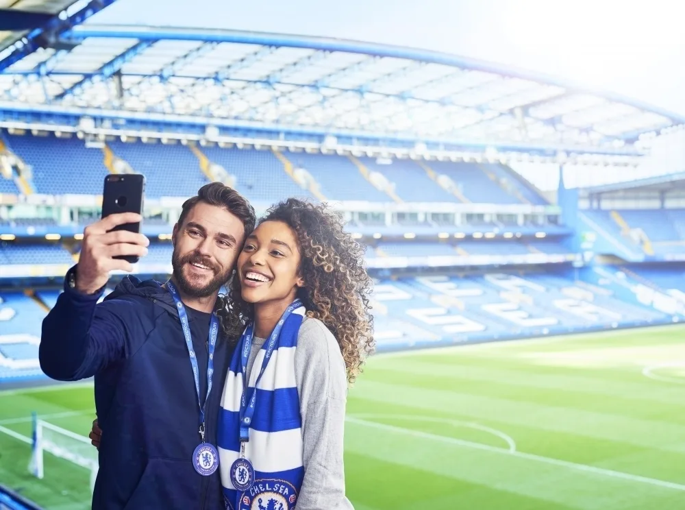A white man with a beard and a Black woman with long hair take a selfie in a football stadium.