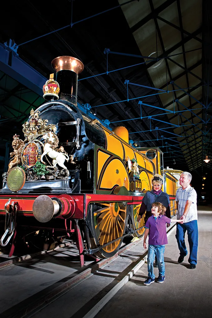 A white man, woman and child standing in front of a steam train in a museum.