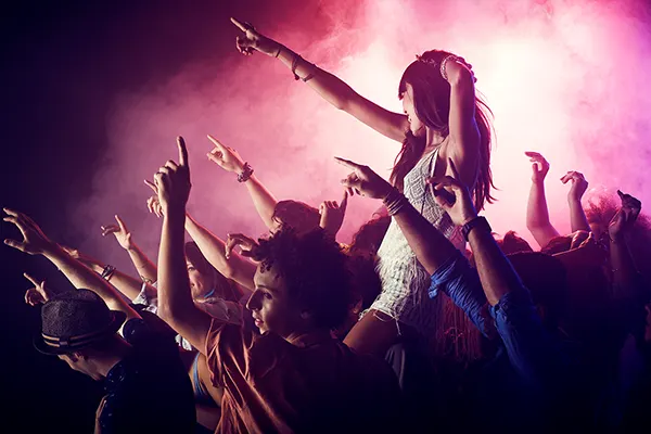 A young woman with long dark hair wearing a white minidress, sitting on someone's shoulders at a gig, she and everyone in the audience have their arms in the air. 