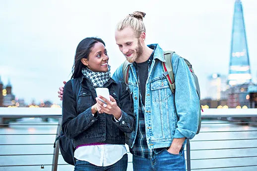 A South Asian woman with long dark hair and white man with a beard and blond hair in a bun smiling and looking at a phone while standing on a bridge over the Thames with the Shard in the background.