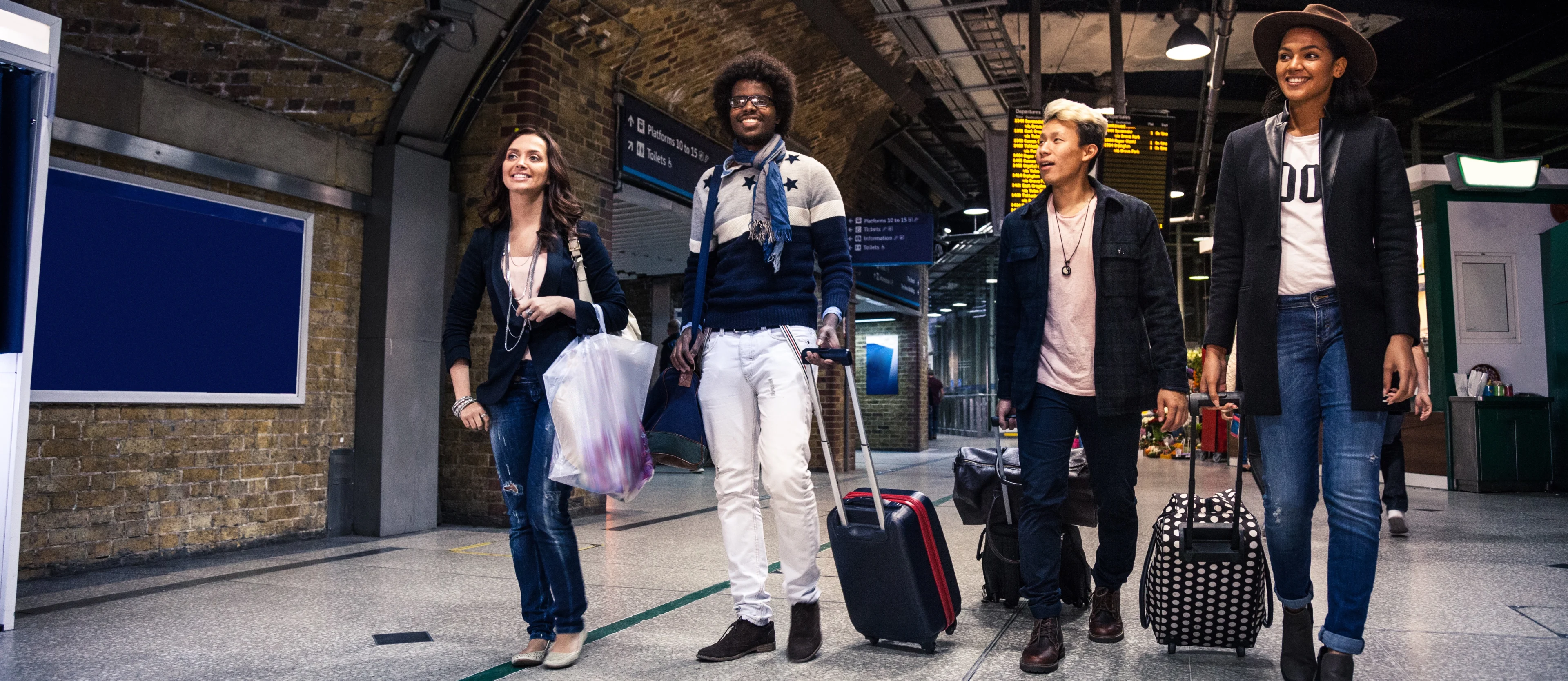 A group of 4 happy friends walk through a train station all pulling or carrying bags and suitcases