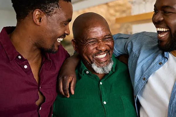 Three Black men laughing together, with an older man in a green shirt embraced by two younger men on either side.