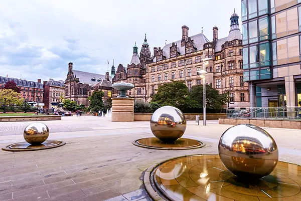 A city square with large brick buildings surrounding it, there is a fountain and statues of large golden balls in the centre.