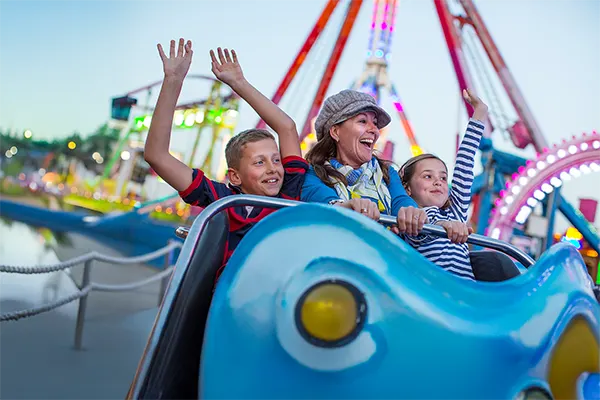 A woman and two children laughing on a blue fairground ride, with colourful lights and attractions in the background.