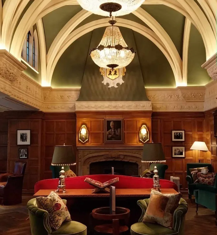 A room with a large high ceiling and chandelier, large fireplace, wood panels and luxurious sofas and chairs. 
