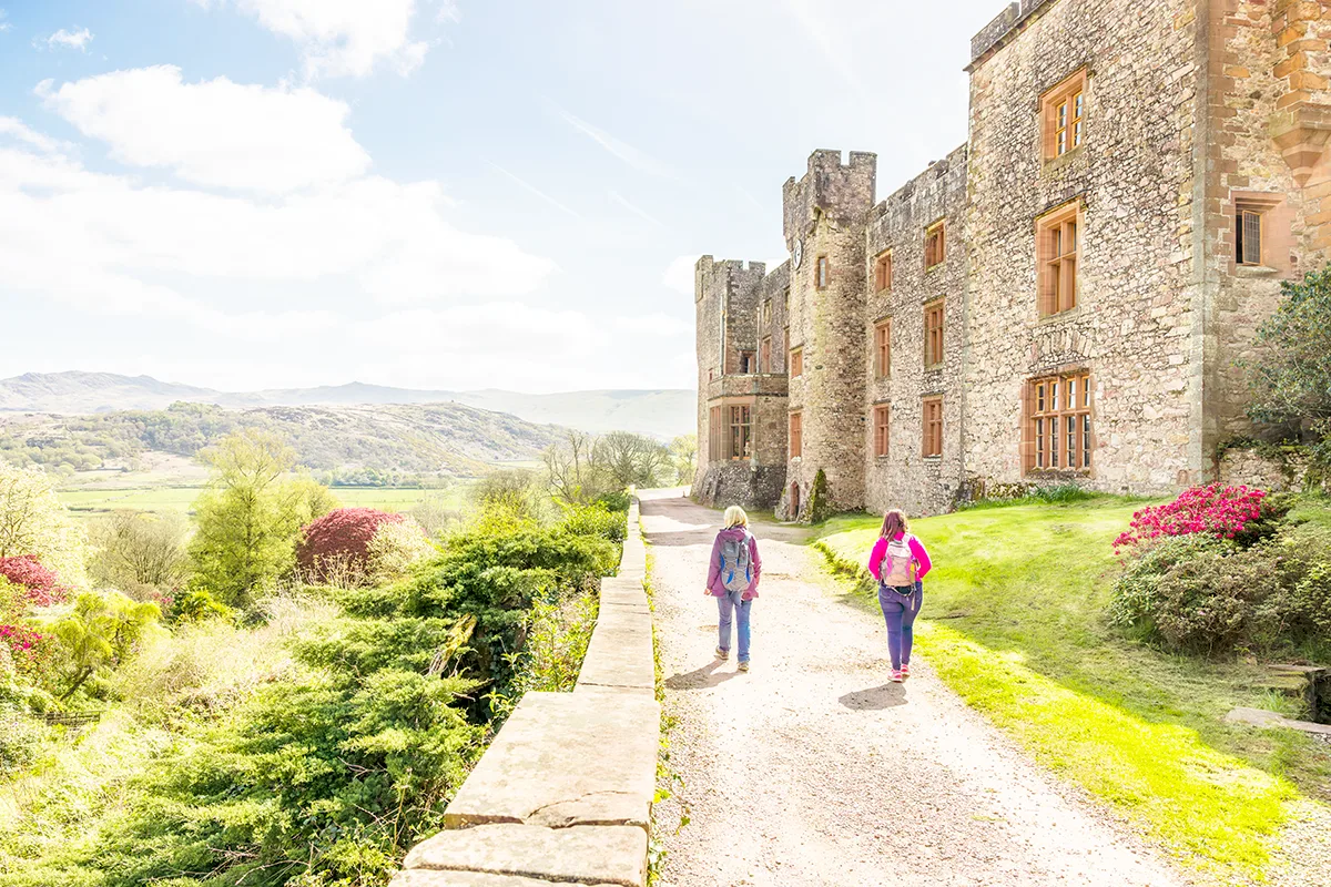 Rear view of 2 women wearing rucksacks walking along a path next to a stone castle on a sunny day. 