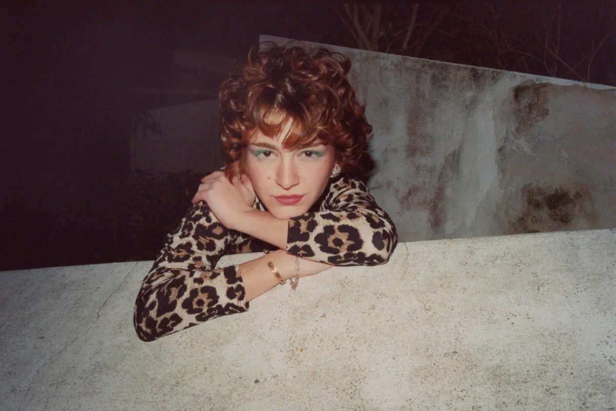 Photgraphic portrait of a person with curly auburn hair wearing leopard print top leaning on concrete surface in dim lighting.