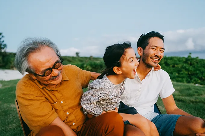 3 generations of an East Asian family – grandad, dad and daughter – are laughing and hugging each other while sitting on green grass.