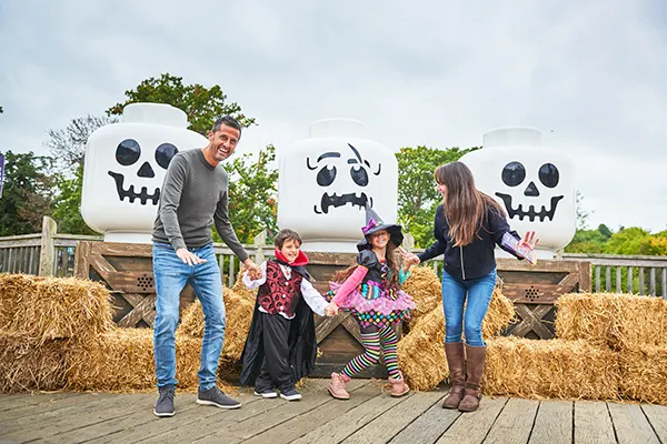 A white family enjoying a Halloween display with children in costumes posing by large LEGO skull decorations on hay bales.