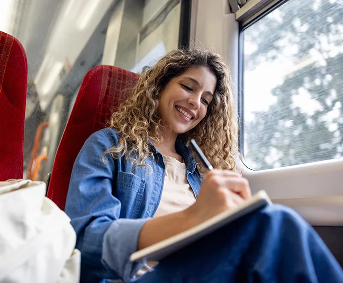 A woman with long blonde curly hair sitting on a train next to a window and smiling as she writes in a notebook. 
