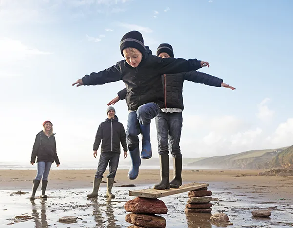 2 children playing on a beach as 2 adults watch nearby. They are all wearing wellington boots and wooly hats. 