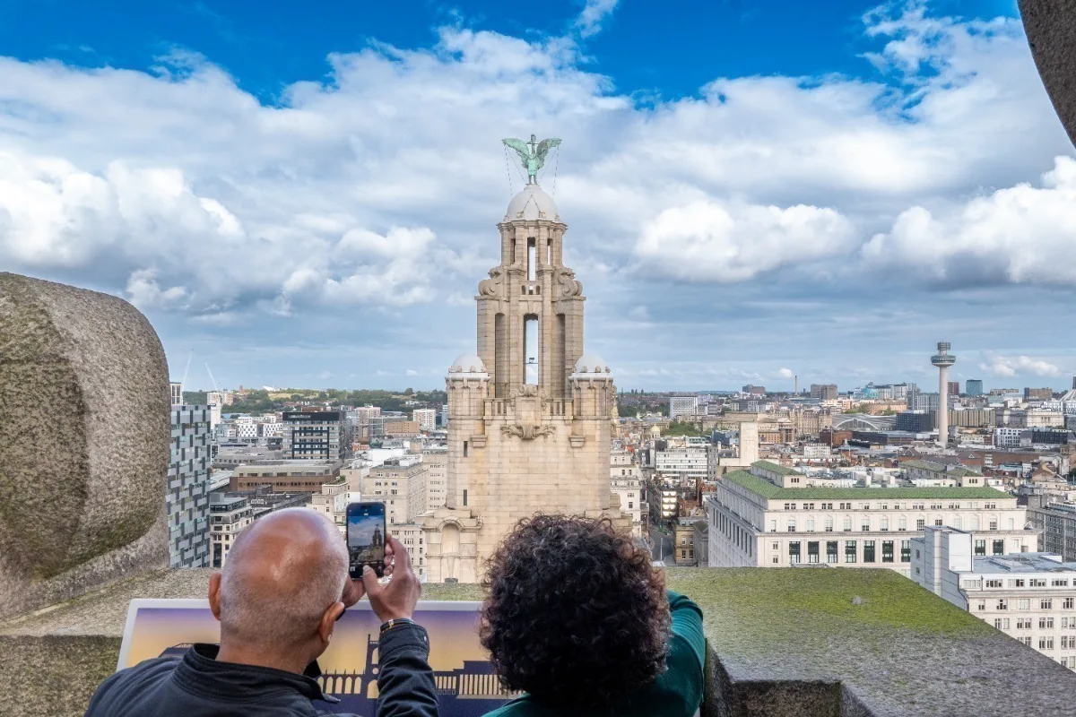 A man and a woman seen from behind, they are at the top of a tall building and looking out at the city skyline to take a photograph.
