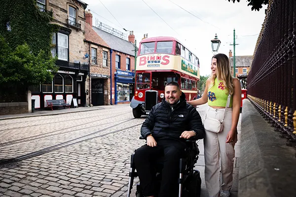 A white man in a wheelchair and a white woman walking next to him on a cobbled street with an old-fashioned red bus in the background. 