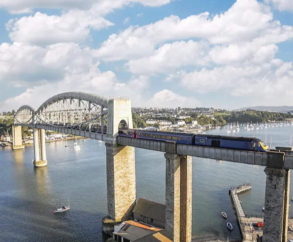 A train crossing a high bridge over a river with small boats in a harbour in the background. 