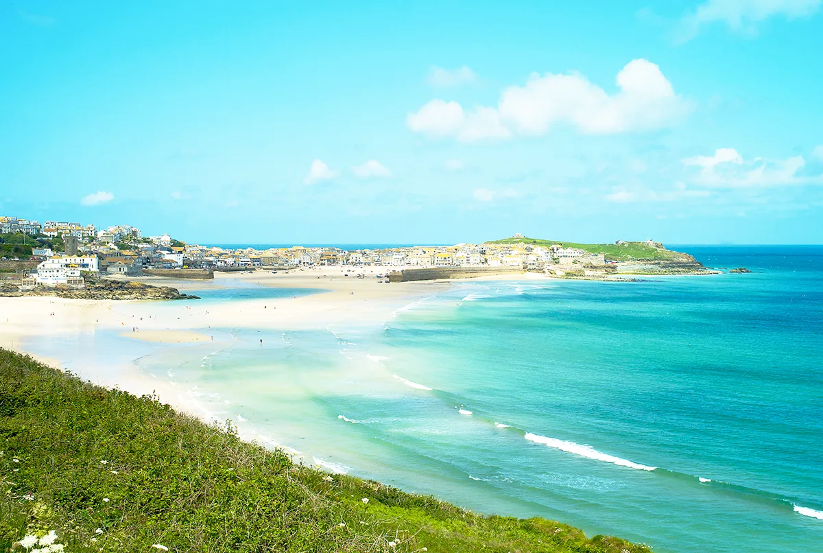 Looking across Carbis Bay in Cornwall on a sunny day, with a sandy beach and bright blue sea and sky, and clifftops in the distance.