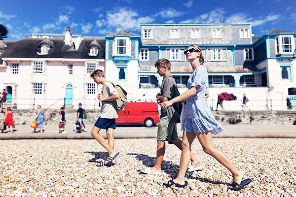 A white woman with blonde hair and 2 blonde teenage boys dressed in summer clothes and walking along a pebble beach on a bright sunny day. 