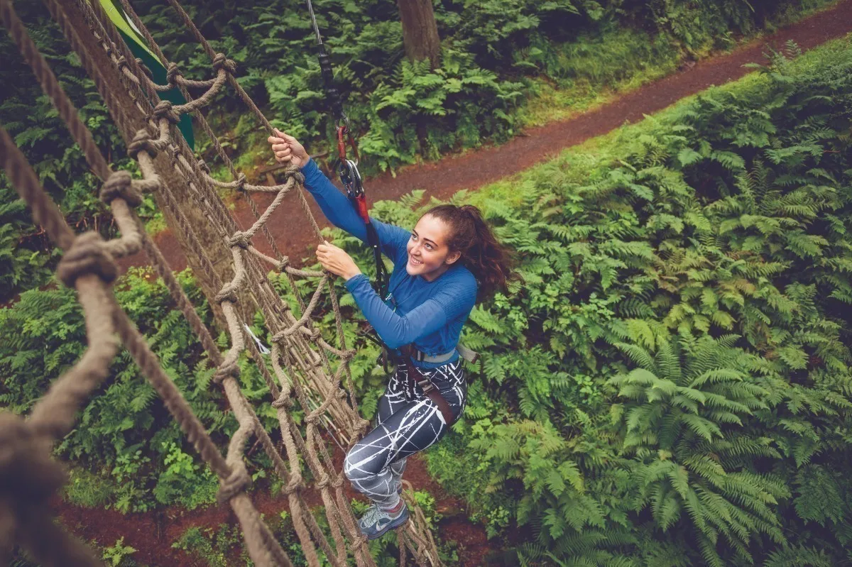 A young woman wearing a harness and hanging from a rope next to a rope climbing wall, high above the ground. 