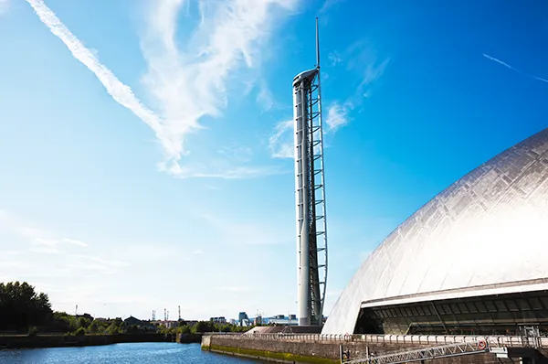 A silver domed building on a riverside, next to a very tall viewing tower. 