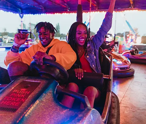 A young Black man and Black woman smiling and punching the air while driving a dodgem at a fairground.