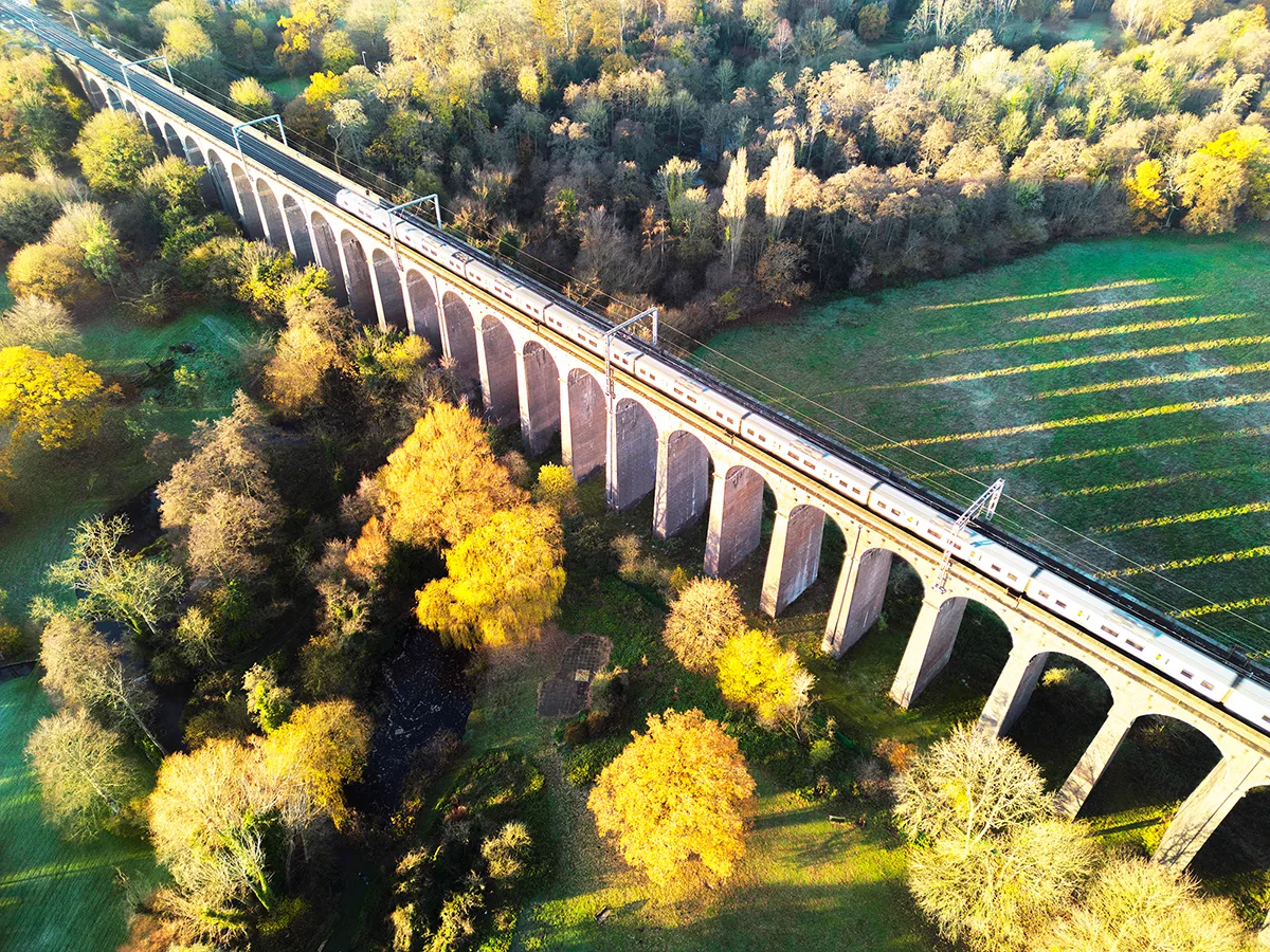 Aerial view of a train crossing a stone viaduct over autumn woodland with golden trees and green fields.