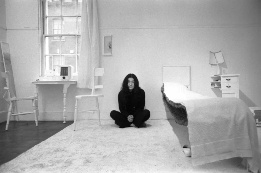 Black and white photo of Yoko Ono (a Japanese woman with long black hair) dressed in black sitting on the floor of a living room that is full of white objects. 