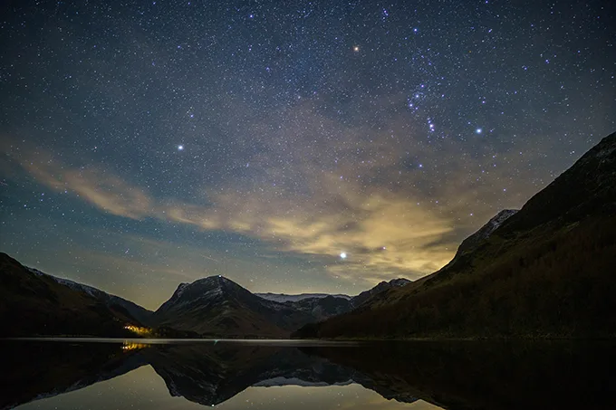 A calm lake surrounded by mountains at night, with a bright starry sky above.