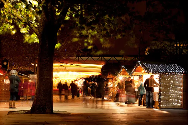 Night-time Christmas market with illuminated wooden stalls and blurred image of shoppers beneath a tree.