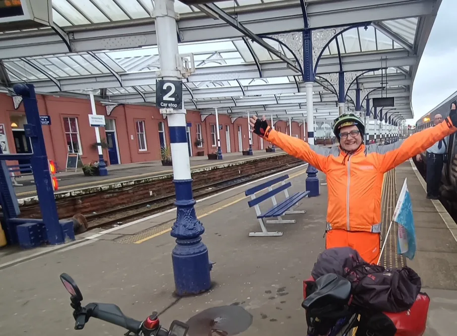 TV presenter Timmy Mallet wearing bright orange clothes and standing on a railway station platform with a bicycle. 