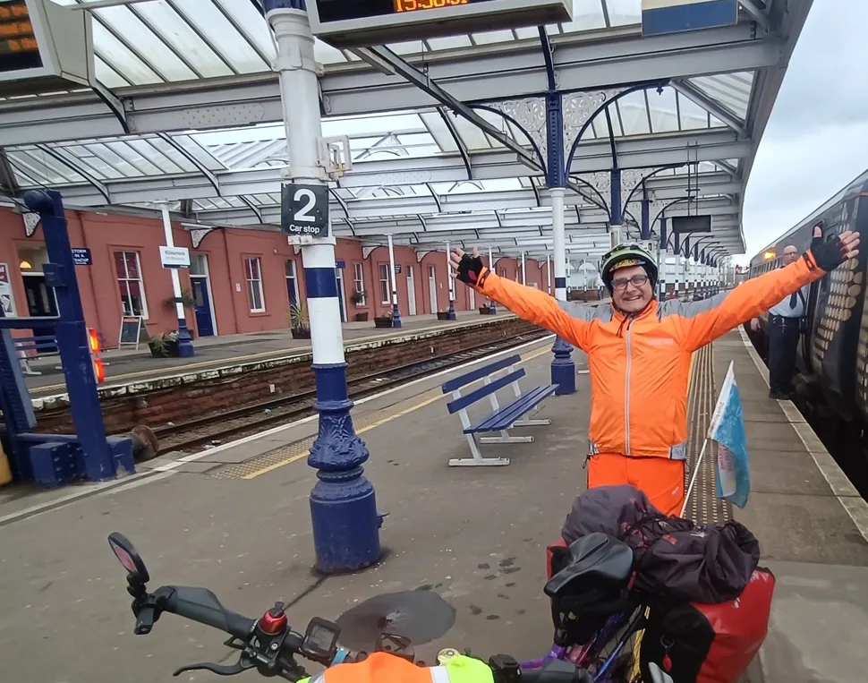 TV presenter Timmy Mallet wearing bright orange clothes and standing on a railway station platform with a bicycle. 