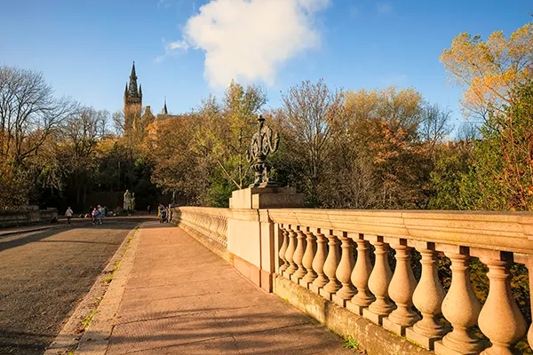 A stone bridge in a leafy park with the spire of a large gothic building visible through the trees in the distance. 