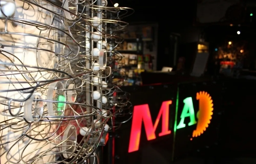 Metal wire sculpture in foreground with colourful "MAD" neon sign visible in the dimly lit interior.