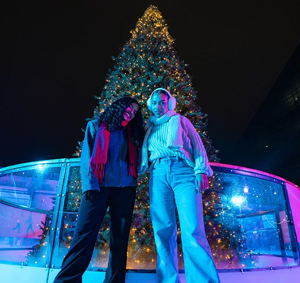 2 young Black women in front of a large Christmas tree at an ice rink.