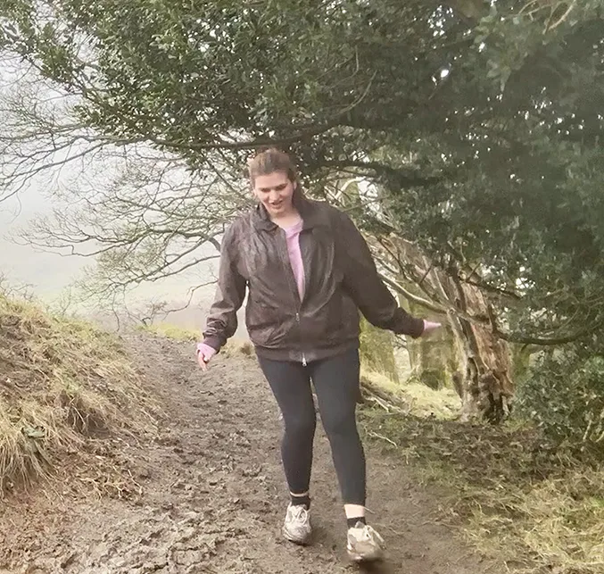 A young white woman with brown hair walking on a muddy path in the country on a rainy day.
