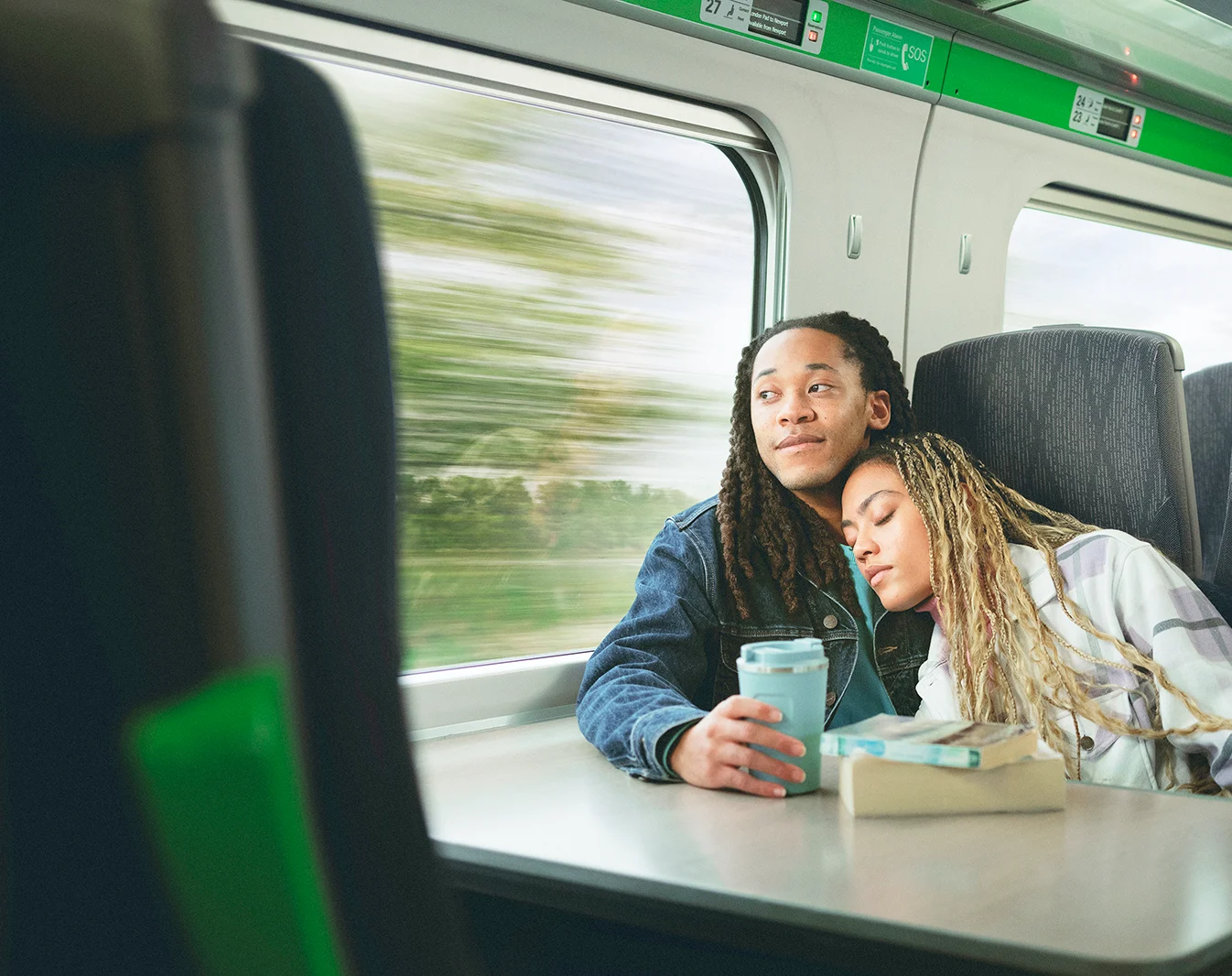 A Black man and Black woman sitting next to each other on a train, he is looking out the window and holding a travel mug while she leans on his shoulder with her eyes closed. 
