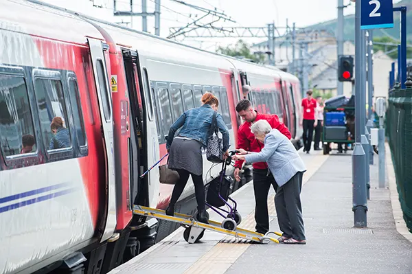 A railway employee helps an elderly woman using a wheeled walker up a ramp onto a train at a station platform, while a passenger guides the walker.