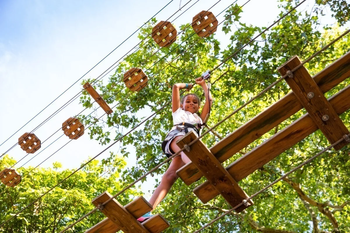 Looking up from the ground at a young Black girl walking across a wood and rope bridge high in the treetops. 
