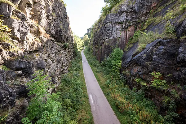 A paved footpath running in the valley between the steep sides of rocky cliffs, dotted with vegetation. 