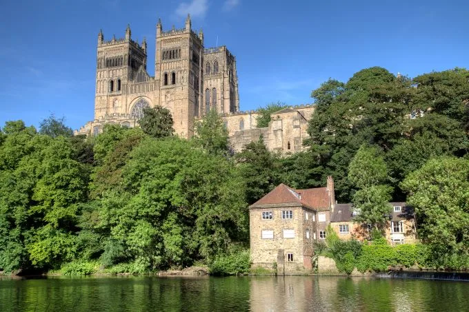 Durham Cathedral sits high upon green trees, meeting the river below. A small building sits on the river below with blue skies overhead.