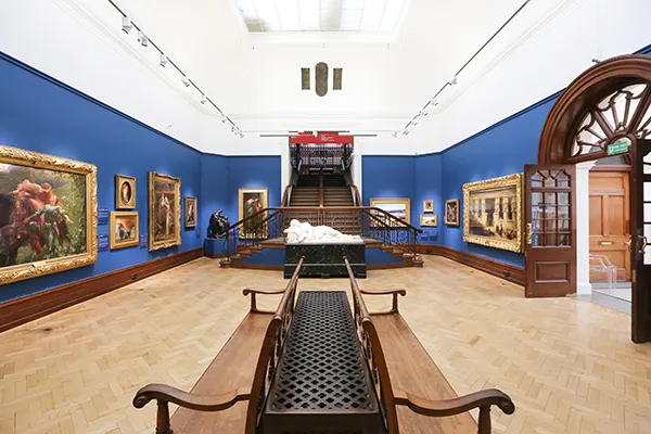 A room in an art gallery with paintings on blue walls, a marble statue on display, and a wooden bench. 