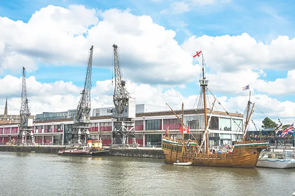 A riverside with cranes and a wooden ship next to a 2-storey industrial building. 