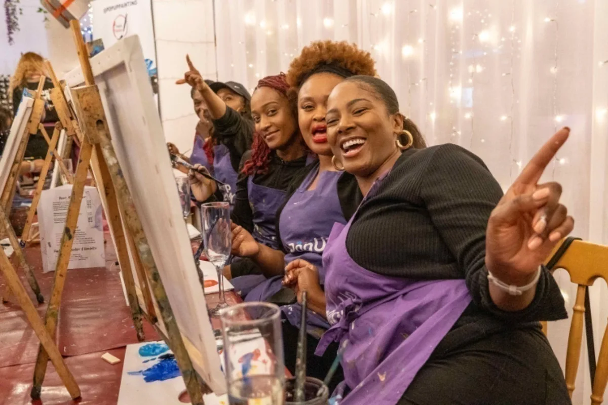 A group of Black women wearing purple aprons and sitting painting at easels, they are smiling and looking at the camera. 
